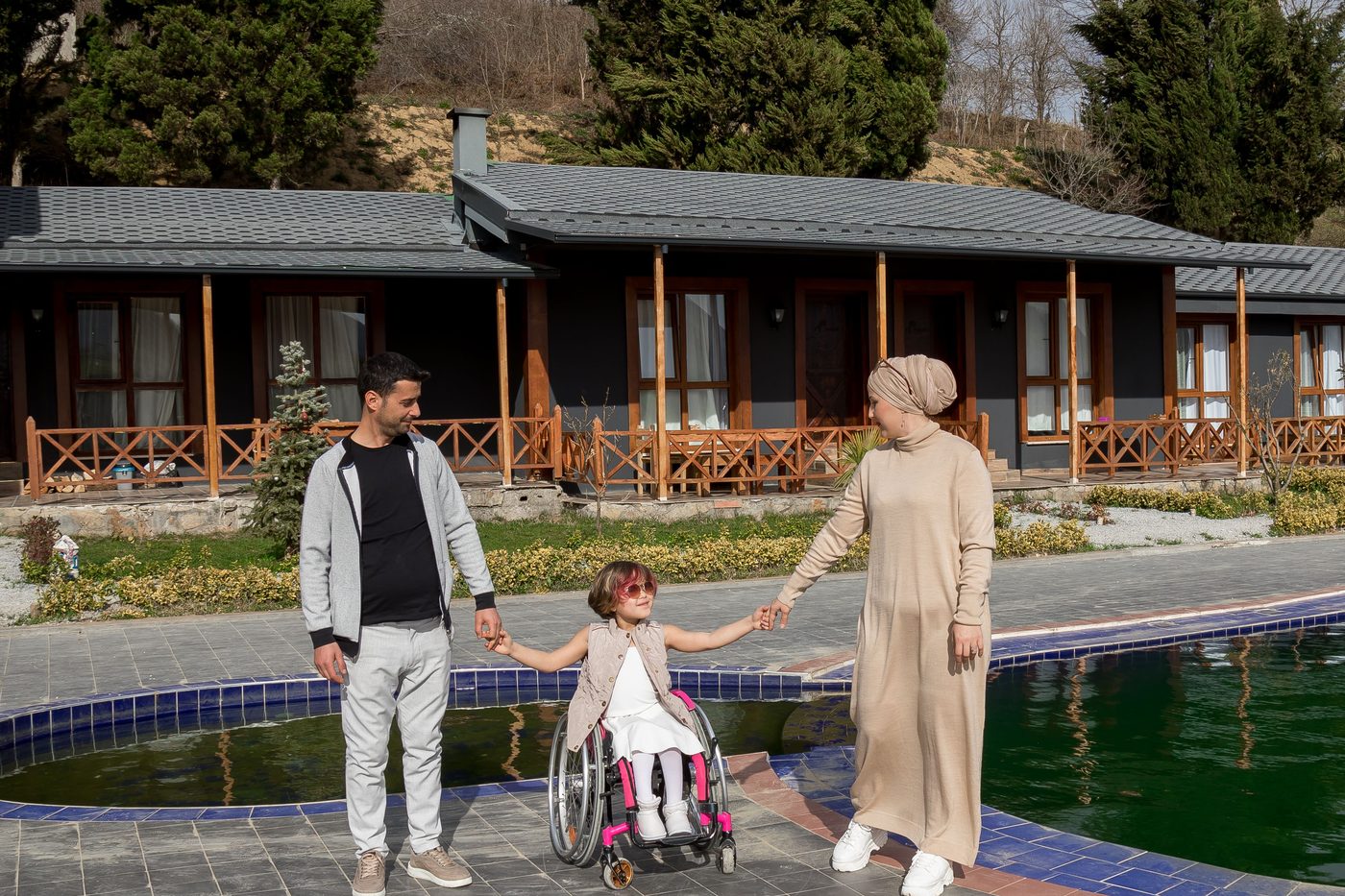 A young girl in a wheelchair holding hands with her parents outside their home.