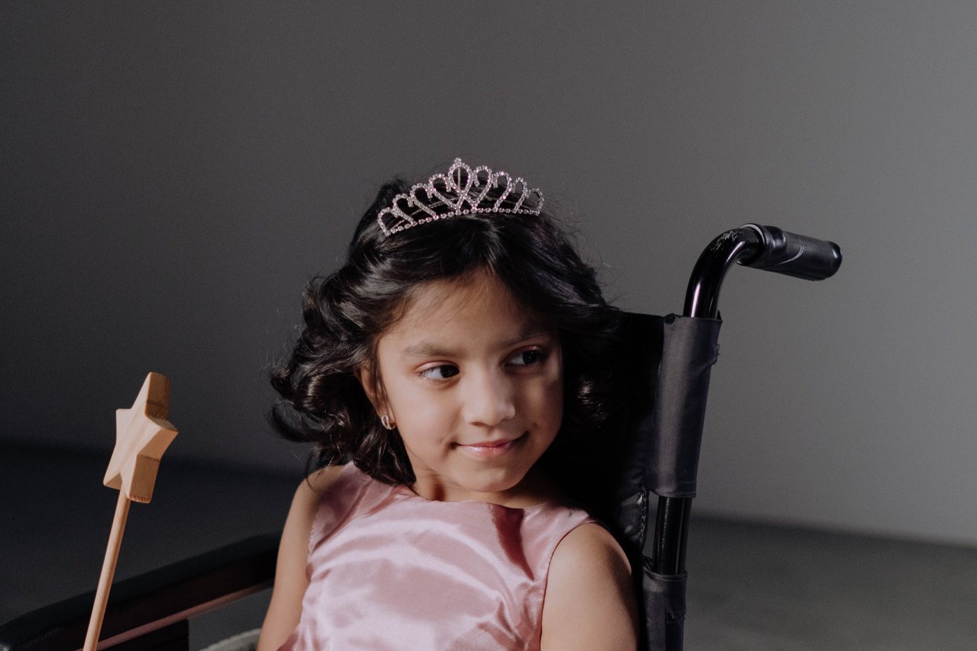 A young girl in a pink dress and tiara, seated in her wheelchair.