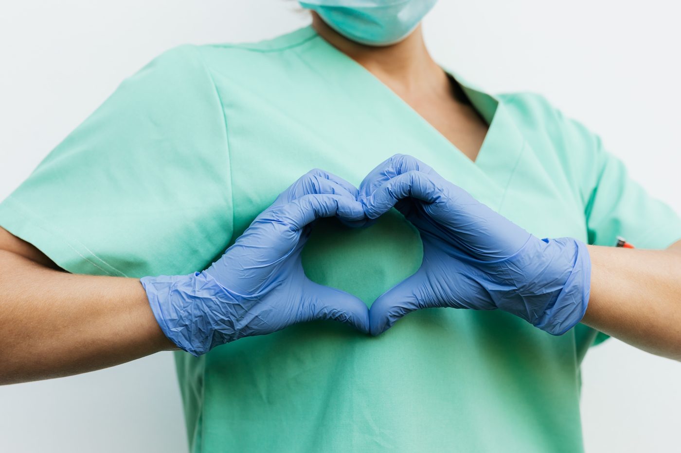 A nurse in scrubs making a heart shape with gloved hands.