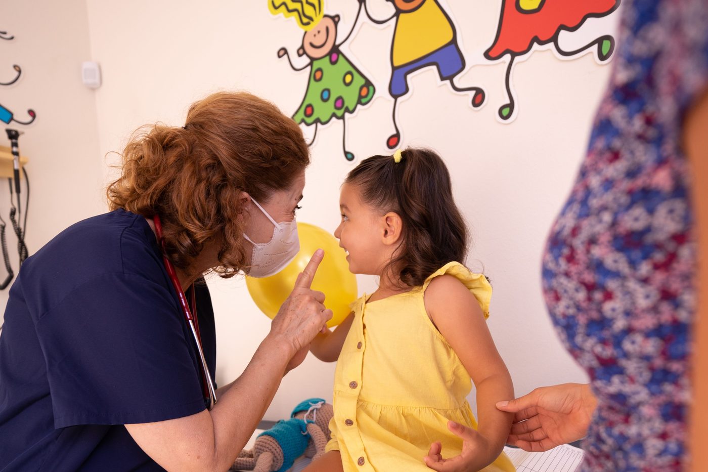 A nurse examining a smiling toddler in a room with cheerful wall art.