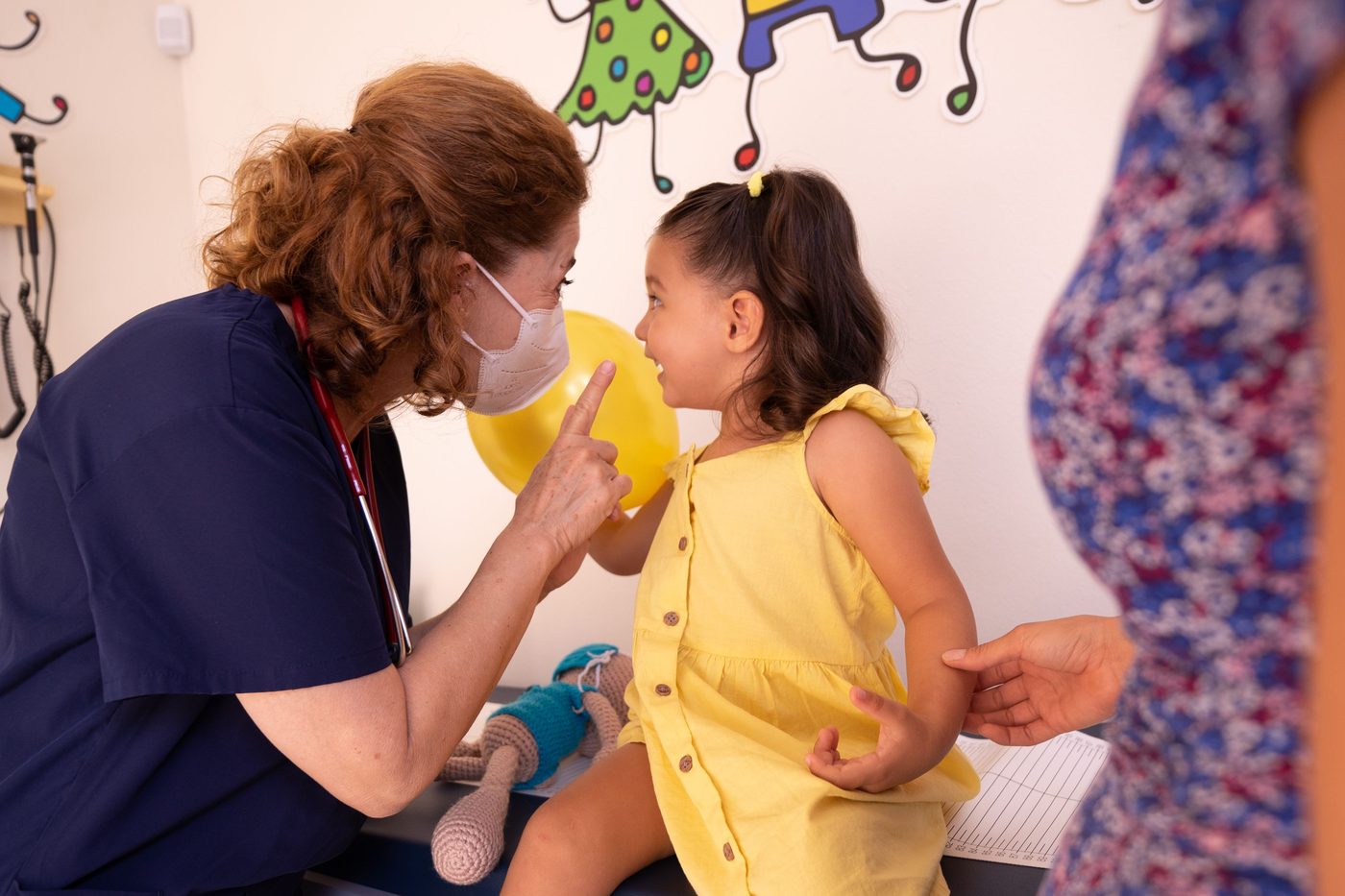 A nurse examining a smiling toddler in a room with cheerful wall art.